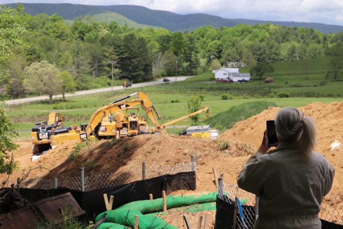 A woman with binoculars looks out on a construction site with heavy machinery and exposed dirt, with green hills in the background.