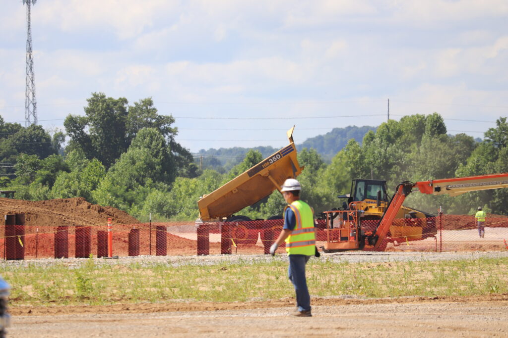 A worker in a yellow vest and a hard hat stands in the middle of a construction site with orange fencing and construction equipment in the background.