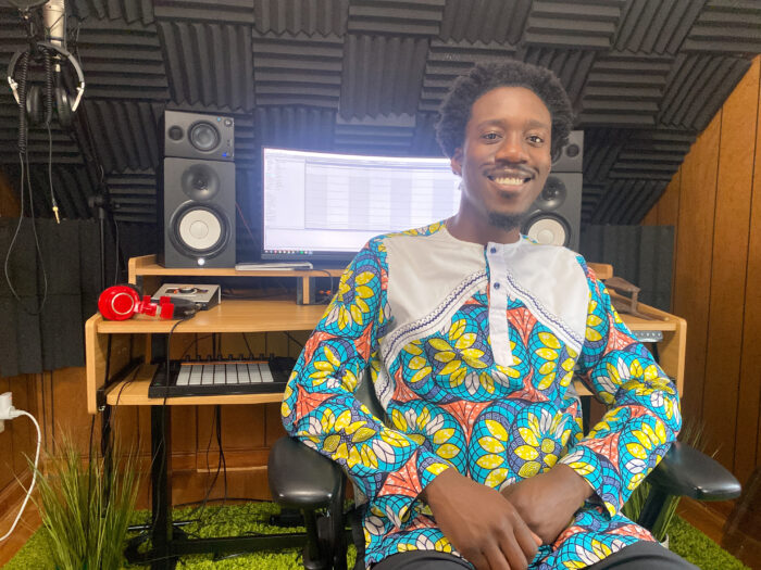 A man smiles for the camera. He leans against a cabinet that had a computer and speakers on top. His jacket is colorful featuring blue, yellow and pink colors.