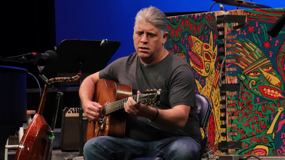 A man with salt and paper hair sits on a chair and plays guitar while singing into a microphone.