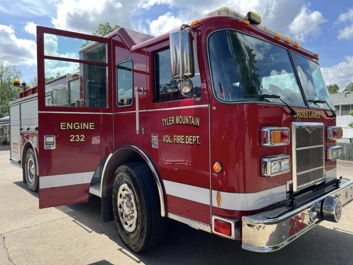 A red fire truck that reads "Tyler Mountain Volunteer Fire Department" and "Engine 232" sits parked on asphalt, with its door open. No one is in the driver's seat, and the sky is bright but partially cloudy overhead. The Tyler Mountain Volunteer Fire Department has helmed emergency response efforts for the Kanawha County town of Cross Lanes since 1951.