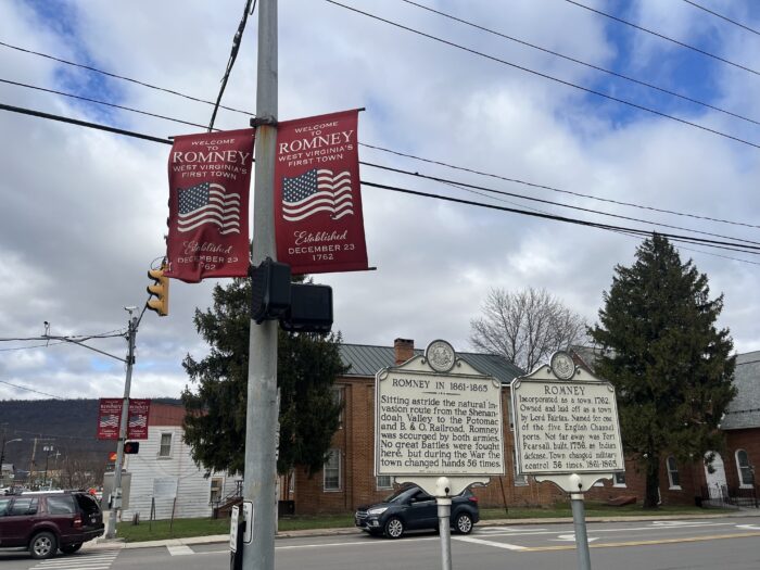 Two banners on a streetlight post read "Welcome to Romney, West Virginia's First Town. Established December 23 1762." The banners are red with a large image of the American flag on them. Behind the lamp post is a town street with cars turning on it. To the right of the banners are two historical markers with additional details about the town's past.