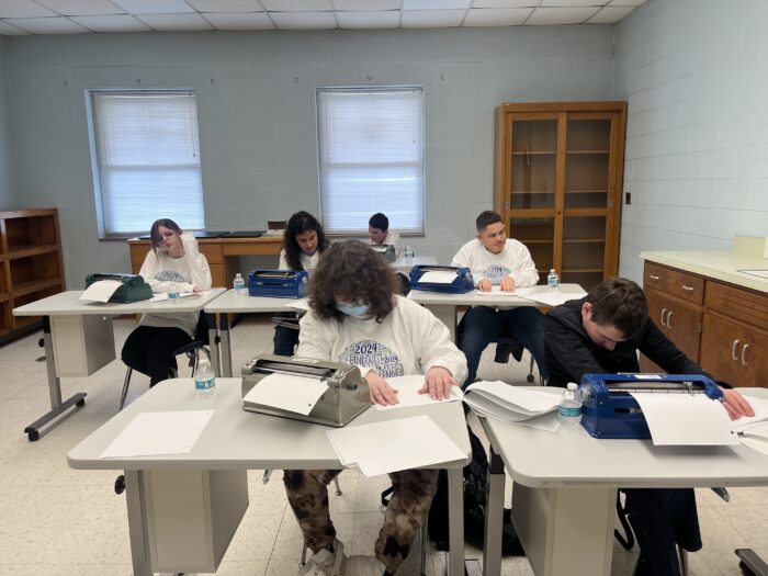 Students seated at run their hands over sheets of paper indented with braille. They appear focused, and have water bottles beside them.