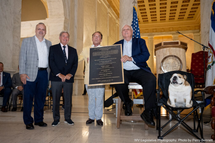 Four men stand arrayed next to an English bulldog seated in a field chair in a marbled, high-ceilinged room with an archway to the left. The man to the left wears a grey checkered jacket over a white shirt and blue pants, while the man next to him wears a navy jacket over a white shirt, blue tie with a yellow circle pattern and dark grey pants. The other two men in frame hold a plaque between them. The man to the left of the plaque wears a pastel yellow jacket over light grey pants, while the man to the right of the plaque, seated on a high stool, wears a navy blue jacket over a white shirt and charcoal pants.