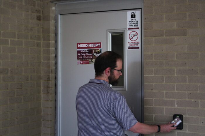 A man in a polo shirt with tight red and white stripes looks to a card swipe to the right of frame as he opens a metal door set into a brick wall. On the door are stickers that read "Safe Weapon Storage Available" and "All Weapons Prohibited"