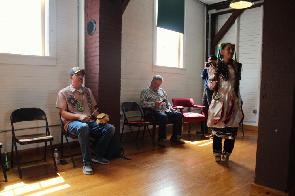 Two men sit on folding chairs against a whitewashed wall below windows with a lot of light pouring through them. The men are playing traditional instruments. To the right of frame a woman dressed in traditional Haudenosaunee clothes dances on a hardwood floor.