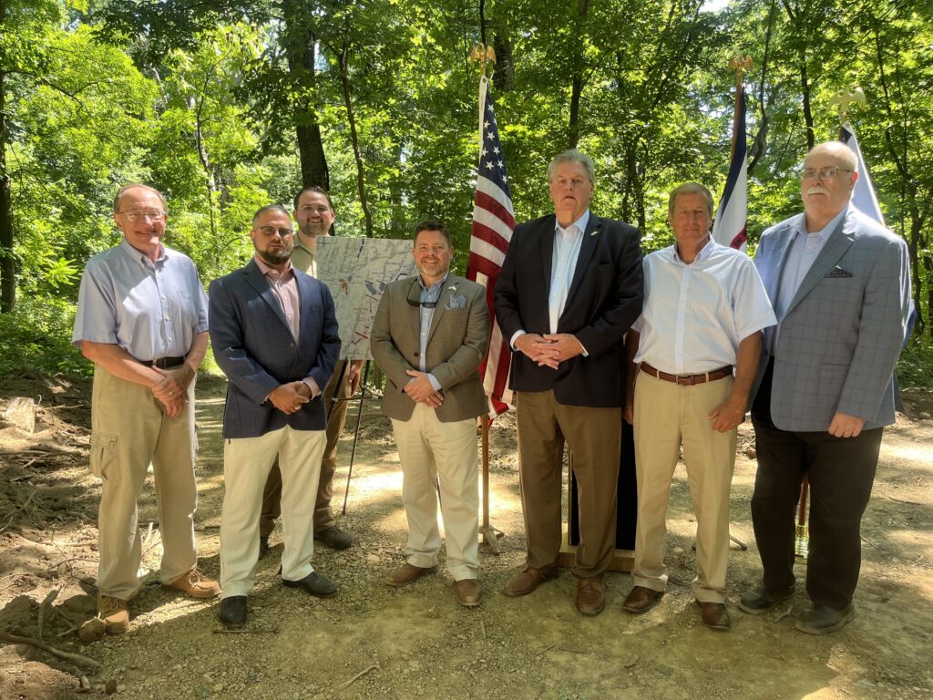 A group of men in formal attire stand on a path in the woods, in front of an American flag and a battlefield map. They are smiling and looking toward the camera.