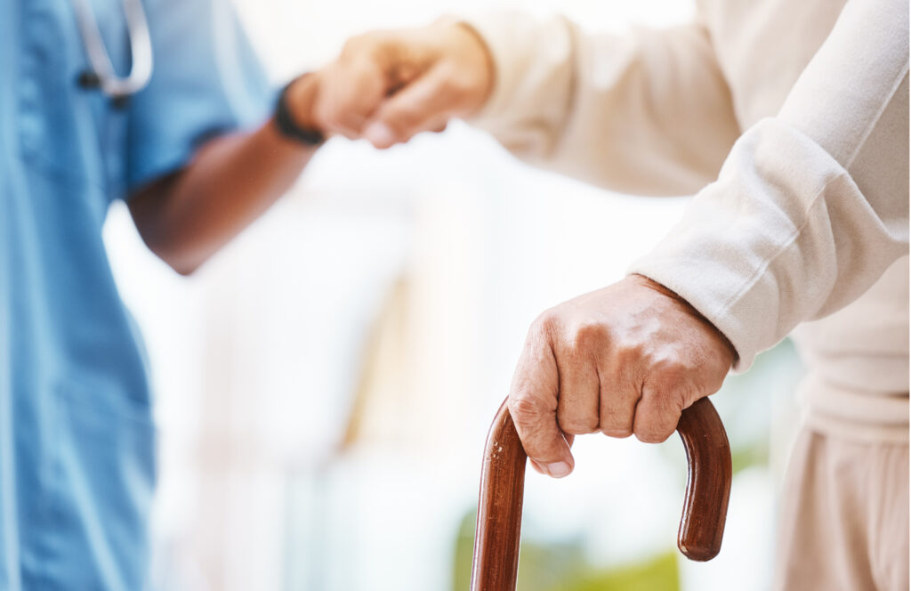 A physician or health care worker dressed in blue scrubs helps an elderly person using a cane to walk with a hand.