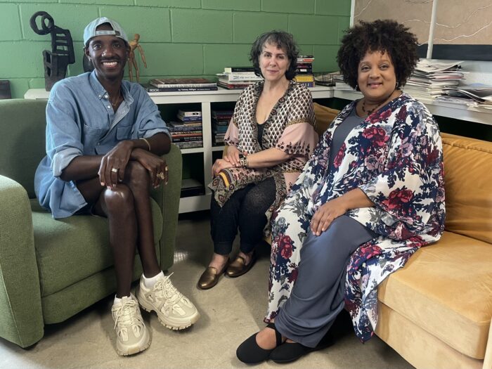 Three people sit on couches and smile toward the camera. Books and magazines are visible atop shelves in the background. A green brick wall is behind them.