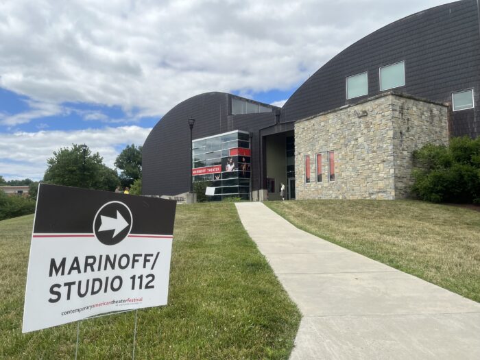 A sign with an arrow pointing to the right reads "Marinoff/Studio 112" and "Contemporary American Theater Festival." To its right, a cement path cuts through trimmed green grass and leads toward a building made of wood panels, stone and glass. The building says "Marinoff Theater" on the front, and the top of the building has a shape resembling the top of a heart.