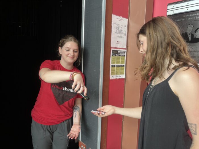 A girl wearing bracelets and a red shirt that says "think theater" with a black heart shape is holding a phone-shaped device that is scanning a barcode on the ticket of a woman waiting in line.