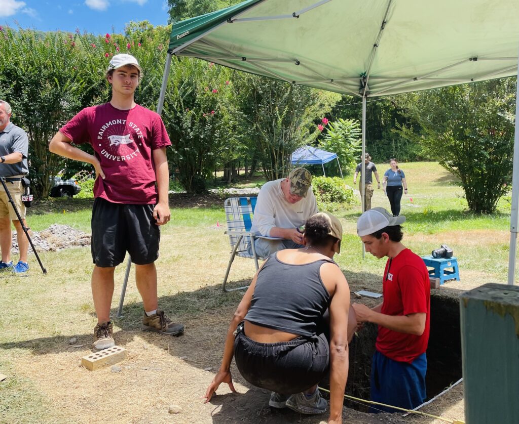 Three people stand under a umbrella. One man is standing waist deep in a whole with a ruler in his hand. A older man sits in a lawn chair with a clip board. A woman crouches beside the whole and is looking a measuring tool A man poses next to the umbrella looking at the camera.