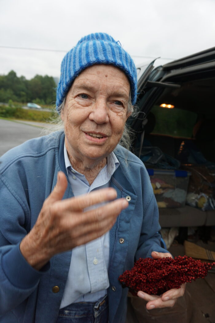 An elderly woman wearing a blue jacket overtop a light blue button up shirt and a blue hat, holds berries in her left hand.