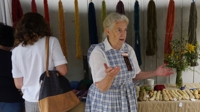 An elderly woman with white hair that is pulled back, wearing a blue and white checkered dress stands in front of a table at an event. Behind her, against the wall, are colorful fabrics.