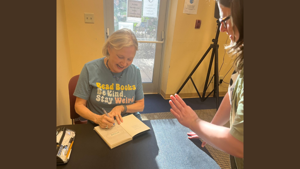 An older woman sits at a table signing books. She is smiling big, laughing as a fan speaks to her.