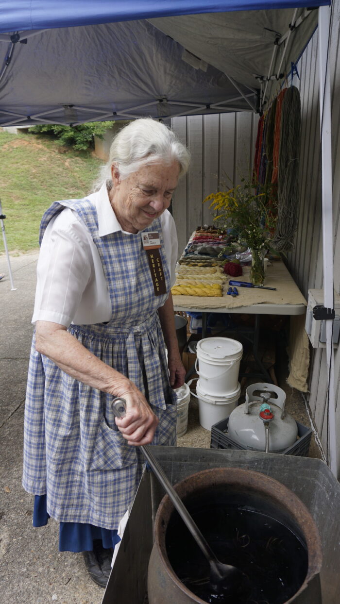 An elderly woman with white hair that is pulled back, wearing a blue and white checkered dress, stirs a pot of dye.