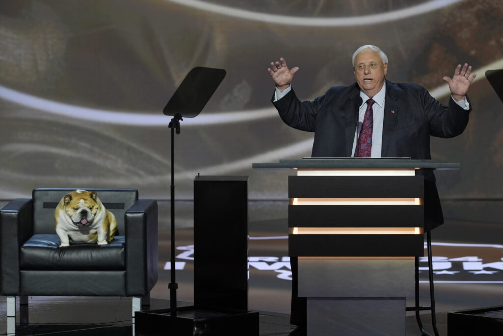 A large man with white hair and wearing a business suit sits behind a podium. Next to him is a brown and white English Bulldog sitting in a black leather chair. Both are sitting on a stage.