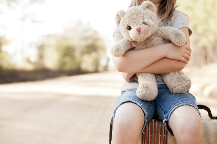 A small child is shown sitting on a suitcase and hugging a tattered teddy bear.