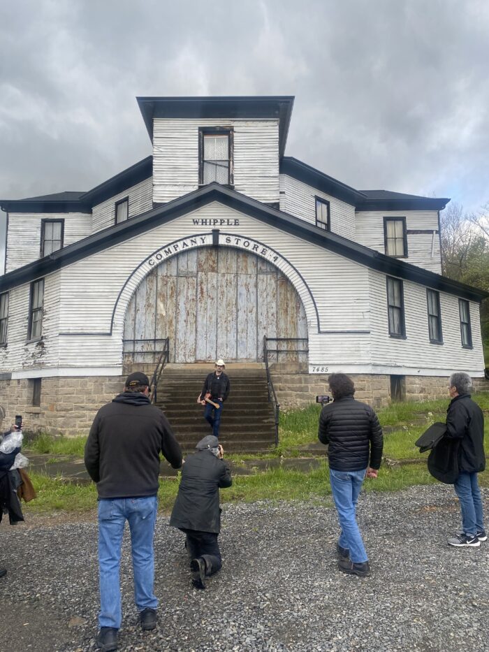 A large, two-tiered, whitewashed wooden structure dominates the frame with dark roof accents and narrow windows dispersed across the facade. The center of the building holds a large arched doorway with a long set of stairs in front of it. Over the arched doorway the words "Whipple Company Store" stand out. On the stairs stands a man holding an acoustic guitar in a white cowboy hat, black jacket and shirt over dark blue jeans. At the foot of the stairs stand an array of people looking at the man on the stairs. One in the center is squatting down to take a picture.