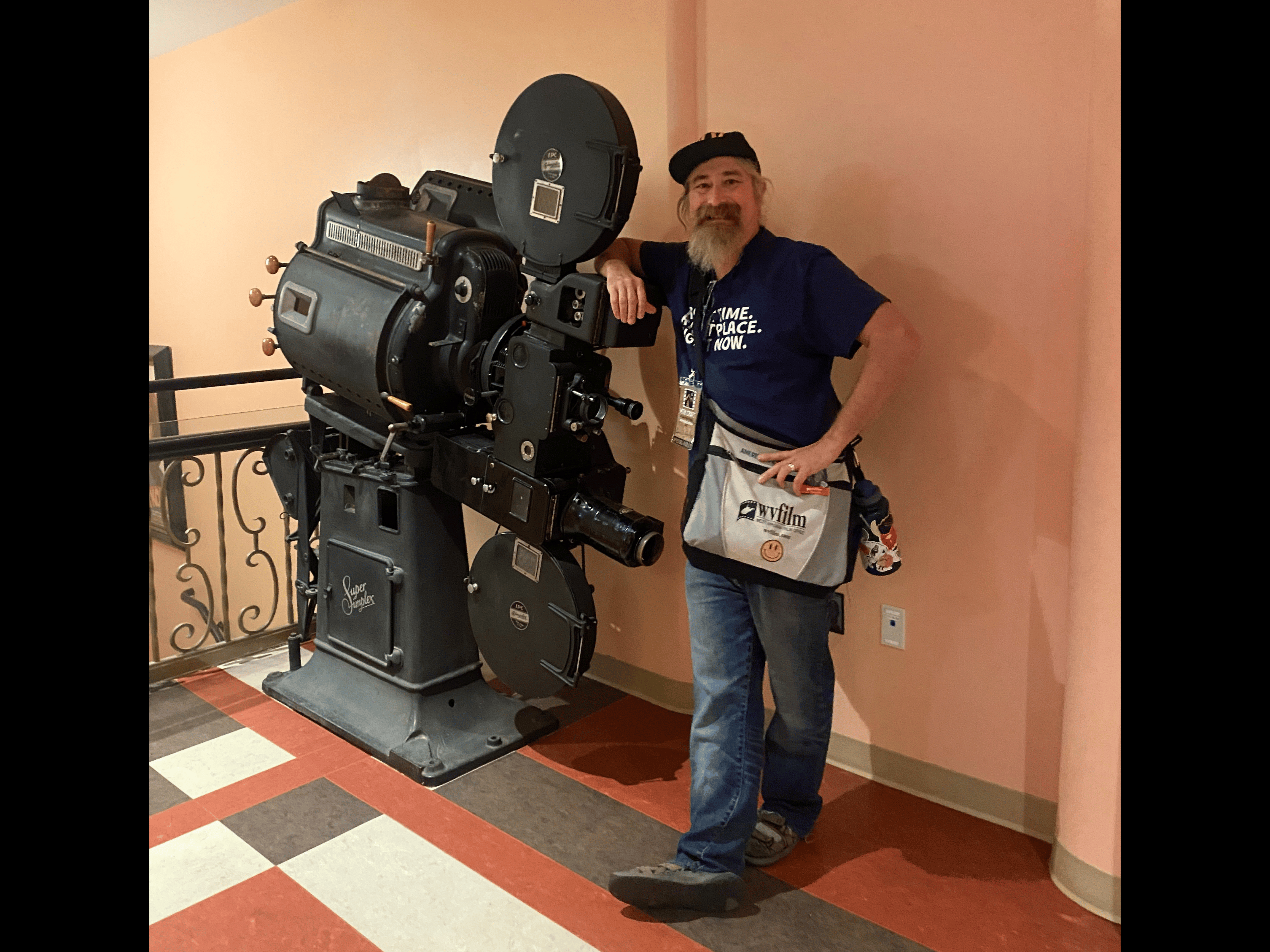 A man stands on a brightly patterned floor of red, black and white tiles as he leans on an old film camera in front of a light pink wall. He sports a white beard and wears a baseball cap with dark blue shirt, jeans and a messenger bag with the WV Film Office logo. The antique camera is positioned in front of a detailed wrought iron railing. The image is bordered by black bars on either side.