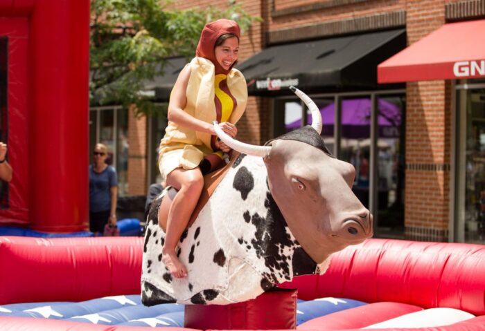 A woman wearing a hot dog costume, including a stripe of mustard up her torso, rides a mechanical bull decorated with a black and white hide. Around her is an inflatable, American-flag themed crashpad. In the far background of the image can be seen storefronts with colorful awnings on a brick-faced building.