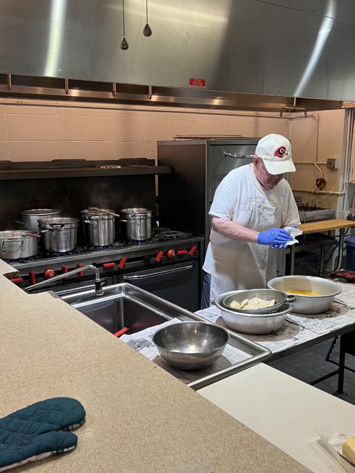 An older man stands over a table in an industrial kitchen. He wears a white ball cap. In front of him are two large bowls. One appears to have uncooked pierogies inside.