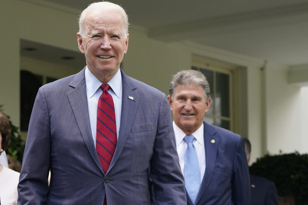 Two older men are seen in a photo walking out of a building. Both are dressed in formal business suits. The man closest to the camera has white hair, while the man walking behind him has gray hair.