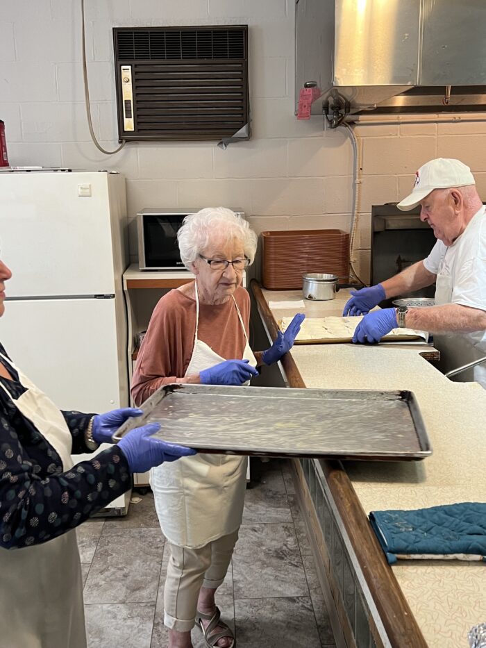 An elderly woman with white hair and wearing glasses stands next to a large baking sheet. She wears a maroon shirt and blue latex gloves.