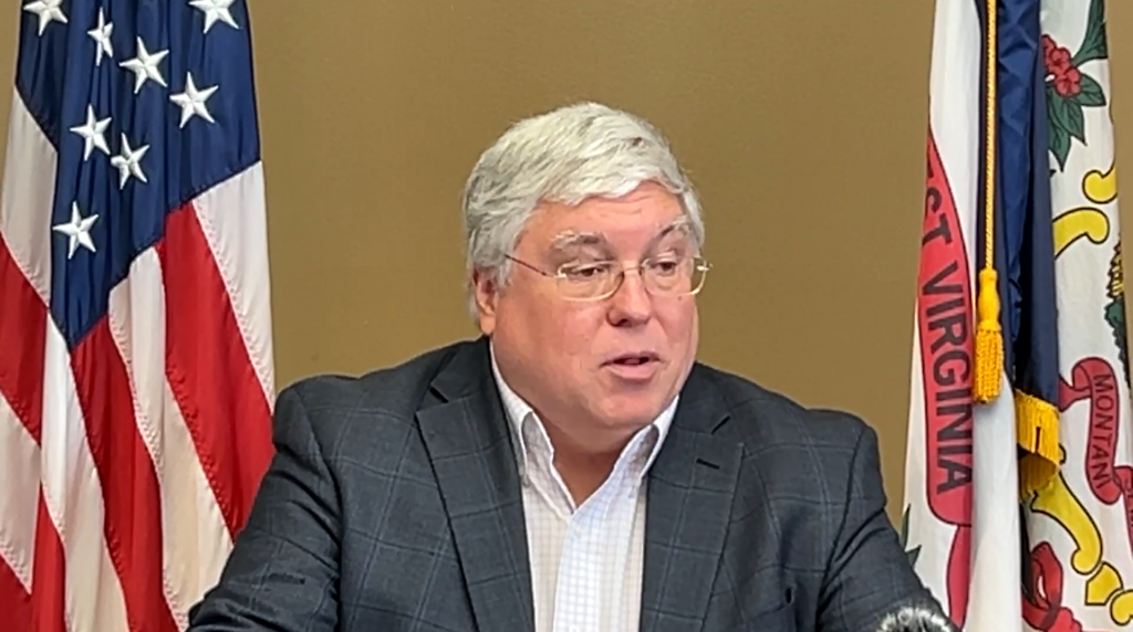 A middle-aged man with white hair and wire rim glasses stands in front of a brown wall in between two flags. One is the United States flag and the other is the West Virginia state flag.