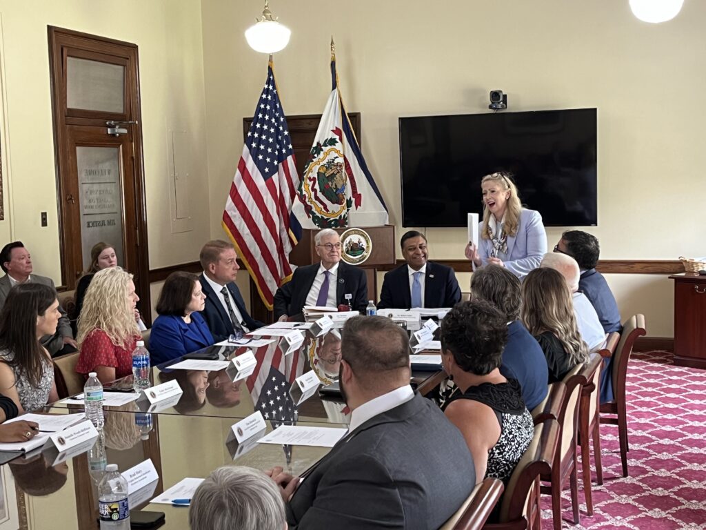 More than ten people are seated at a long wooden conference room table, listening to a woman wearing a blue blazer and pants speak. The carpet in the room is red and the American flag and West Virginia state flags are visible in the background.