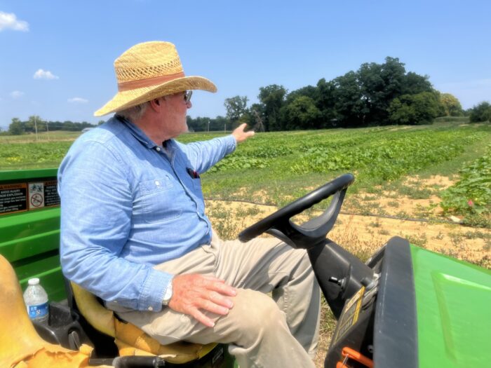 A man in a blue, collared shirt and straw hat sits on a green cart and gestures toward a field of plants growing low to the ground. The earth looks slightly dry, and lines of small, black tubes run alongside the crops.