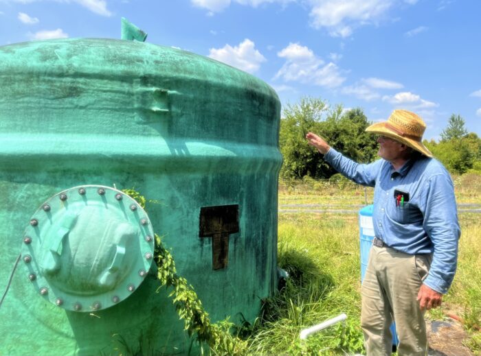 A man in a blue, collared shirt and straw hat gestures to a large, turquoise water tank in the middle of a field.