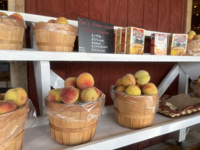 Baskets of peaches sit atop white shelves outside of a wood-plank building painted red.