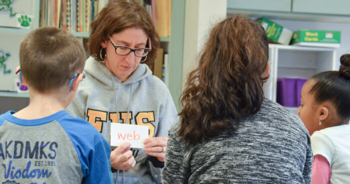 A female teacher shows a flash card to three students who are learning to read. The card reads, "Web."