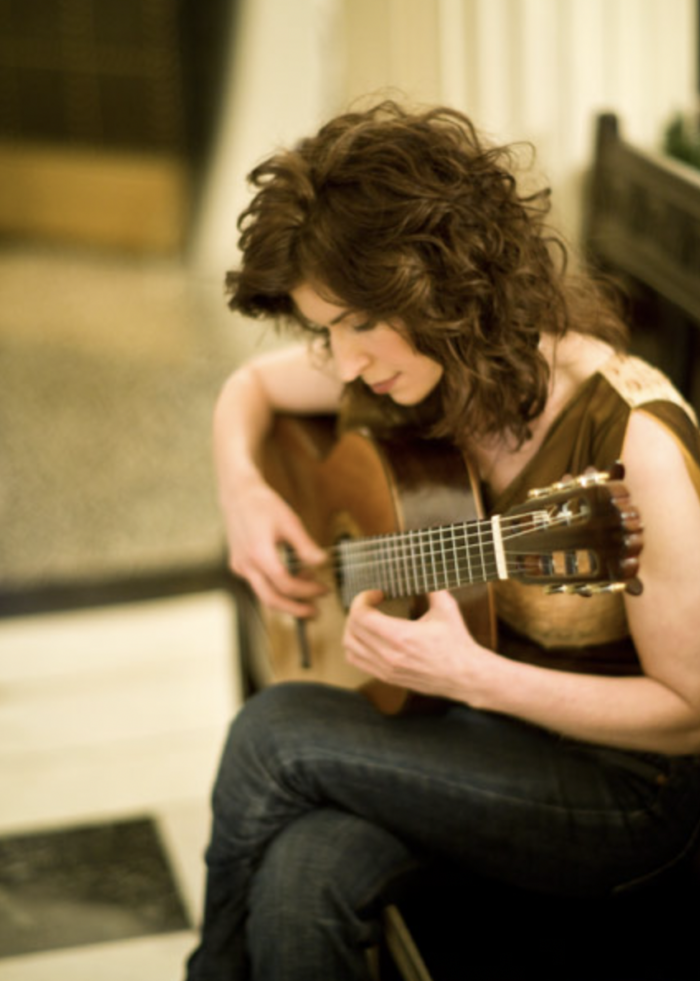 A woman with curly brown hair sits with her legs crossed playing a guitar on her lap