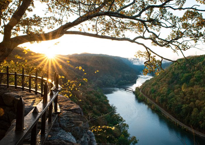 The sun sets over a river cutting through a mountainside. Trees covered in green leaves line each side. On the left-hand side of the photo, an overlook platform for tourists is visibile.