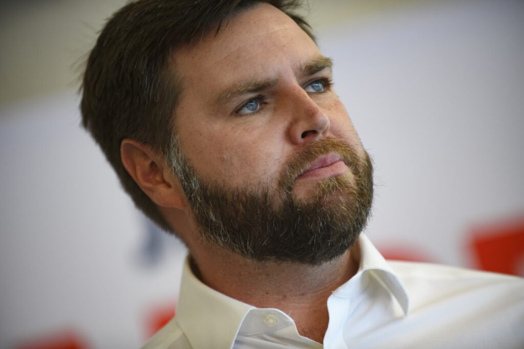 A close up of an older millennial man with brown hair and beard. He has blue eyes and looks off the camera, not smiling.