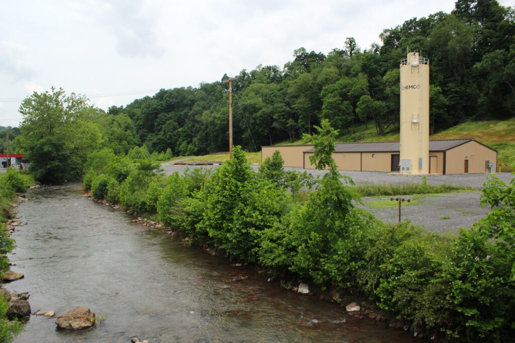 A waterway runs on the left of frame with a tuft of vegetation to its right, separating the water from a parking lot. In the background, a large tan tower with "Chemco" branding on it dominates a low-slung building at the base of a forested slope. A cloudy sky dominates the rest of frame above.