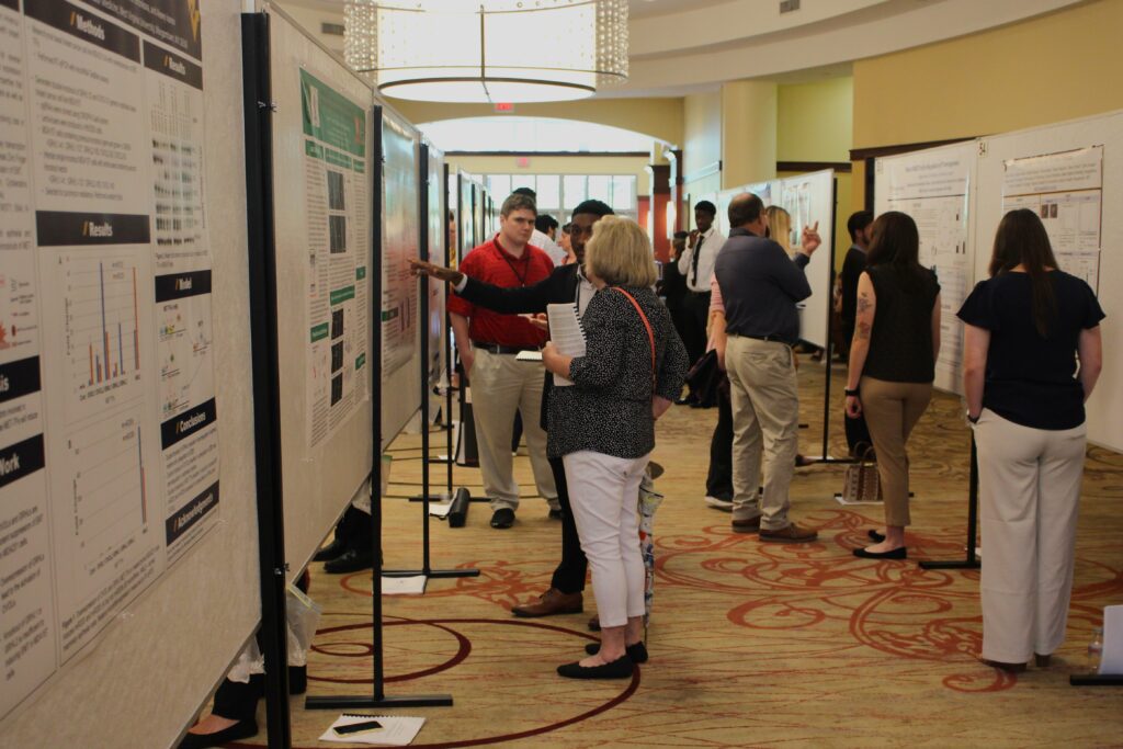 Large informational posters are arrayed on tall stands to form a narrow passageway inside of a larger space with yellow carpet and walls and a white ceiling with large chandeliers. Between the poster walls stand about a dozen or more people looking at the posters, most facing away from the camera.