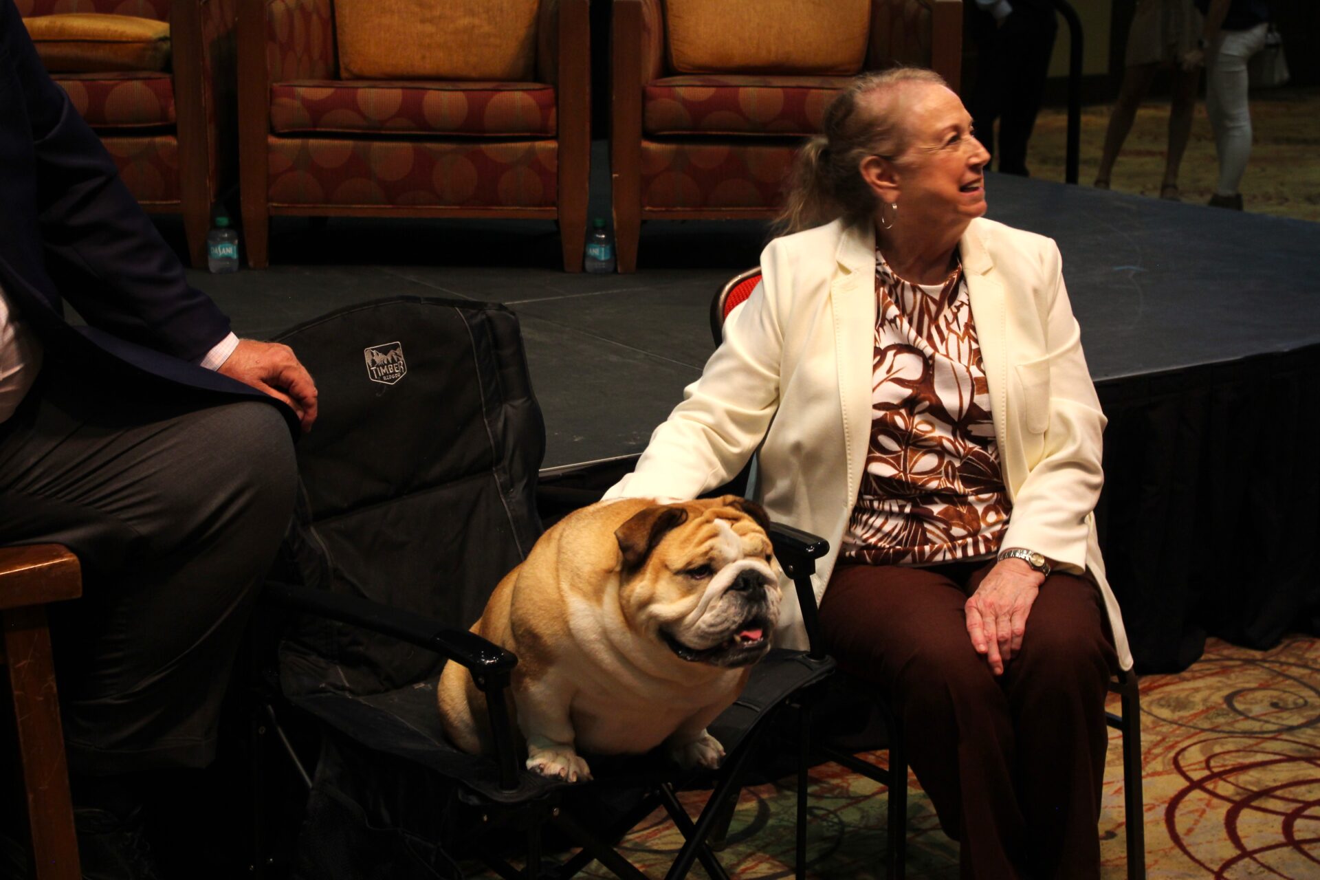 A portion of a man in a dark suit can be seen, primarily his hand, arm and leg, seated to the left of frame. In center of frame is an English bulldog perched on a chair. Next to the dog sits a woman wearing a white blazer over maroon pants and a maroon floral print blouse. She looks to the right of frame. Behind them, on a short stage, are visible brown lounge chairs.