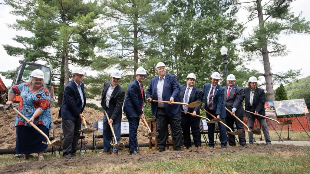 Several men and one women wear formal attire and hard hats. They are lined up and holding shovels in front of a pile of dirt. They are motioning toward the ground, and scooping small bits of dirt into their shovels. Behind them sits a row of chairs, and several posters depicting 3D renderings of a new exhibit at the West Virginia State Museum.