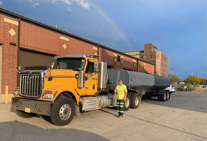 A truck carrying a long water tank sits outside a building, parked in atop asphalt. A man stands beside the vehicle.