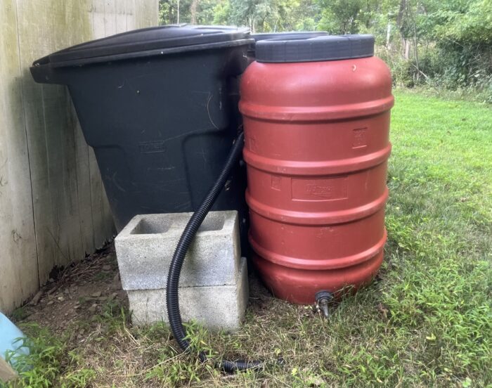 A red barrel with a faucet on the bottom and an opening on the top sits beside a black plastic trash can and a stack of two cinderblocks in a yard.