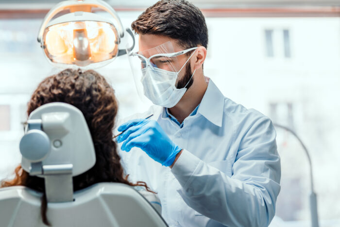 A patient is seen sitting in a dentist's chair from behind. We can see the dentist's smile behind his respiratory mask and shield glasses. He is wearing blue gloves and is holding a metal instrument.