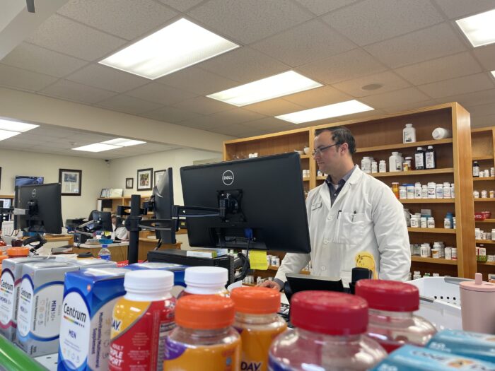 a man in a white coat looks a monitor. In the forefront of the photo are supplement bottles. He is behing a counter, at a pharmacy, with wooden shelves filled with pill bottles behind him.