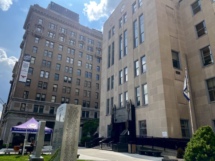 Two buildings stand tall against a bright, partially cloudy sky. They have windows on every floor. In the foreground sits an upright commemorative plaque, a pop-up tent with an individual seated beneath it and the West Virginia flag. The building closest to the camera reads "Harrison County Courthouse," with two decorative eagle sculptures displayed on either side of the sign.