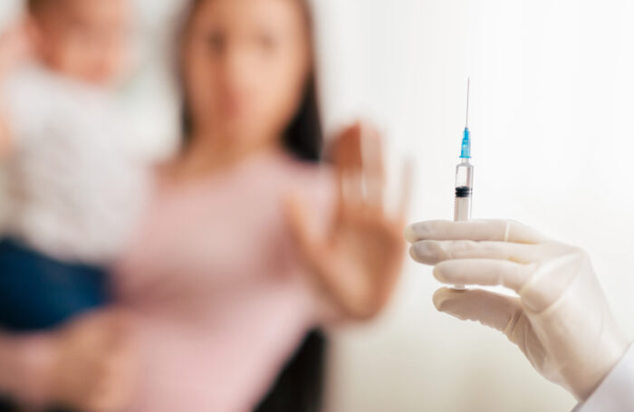 In a blurred background, a brunette woman wearing a pink shirt and holding a child wearing a white shirt and jeans holds up her other hand in a "stop" signal to a white-gloved hand in the foreground holding a syringe.