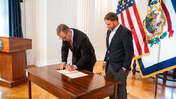 Two men in suits stand before a polished wooden table, with the American and West Virginia flags behind them. The man on the left is signing a sheet of paper atop the table, and the man on the right is watching him do so, pen in hand, awaiting his turn to sign.
