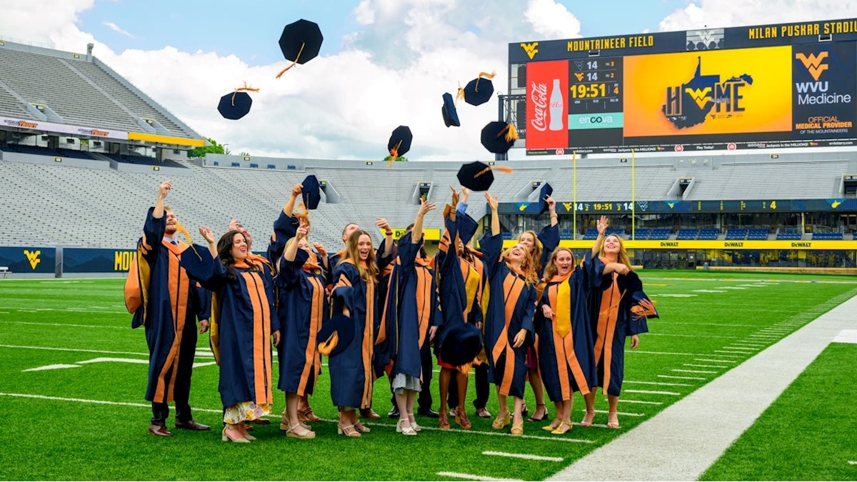 A group of graduates wearing black graduations gowns and throwing their caps stand on a football field.