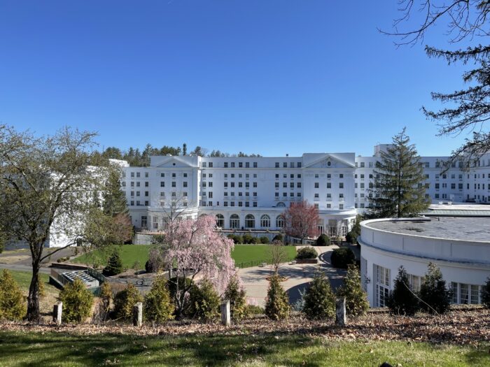 a white hotel with many windows sits below a small hill. There are minimally blooming trees. Its early spring and a sunny but cold day.